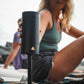 Woman sitting on beach holding a black portable speaker with a stake attachment underneath, she is staking it on the sand.
